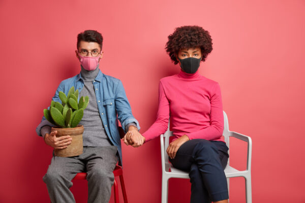Infected married woman and man have corona virus wear protective masks and hold hands sit on chairs and stay at home during self isolation or pandemic isolated on pink background. Quarantine time