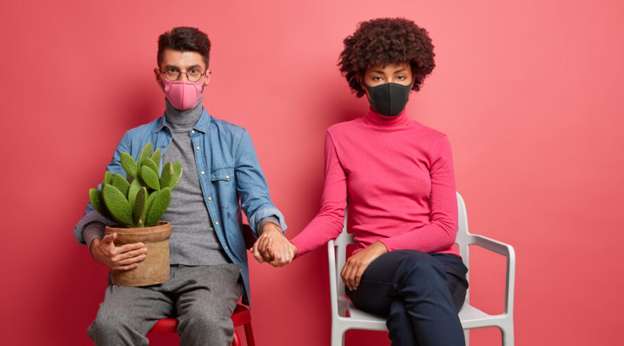 Infected married woman and man have corona virus wear protective masks and hold hands sit on chairs and stay at home during self isolation or pandemic isolated on pink background. Quarantine time